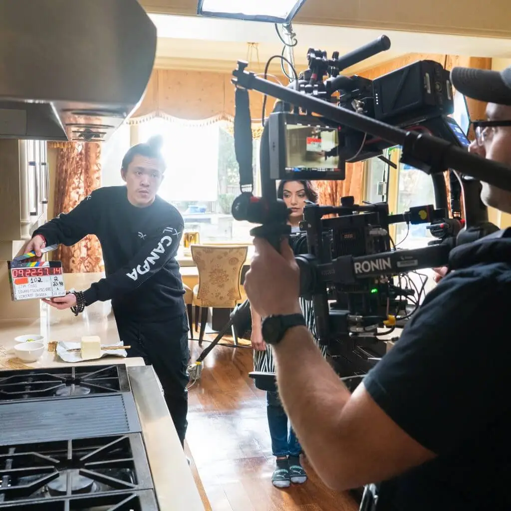 FX Productions Canada A Toronto Video Production crew films a scene in a kitchen, with a camera operator capturing a man holding a clapperboard and a woman standing beside him. Toronto Video Production Company