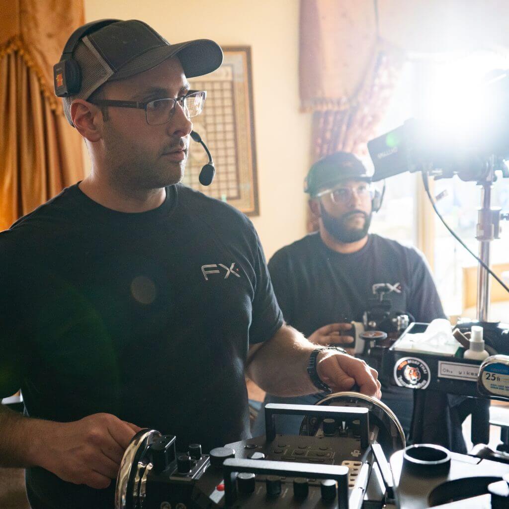 FX Productions Canada Two crew members in FX Productions Canada shirts operate film equipment and cameras indoors, sunlight streaming through a window behind them, showcasing the expertise of this leading Toronto Video Production company. Toronto Video Production Company