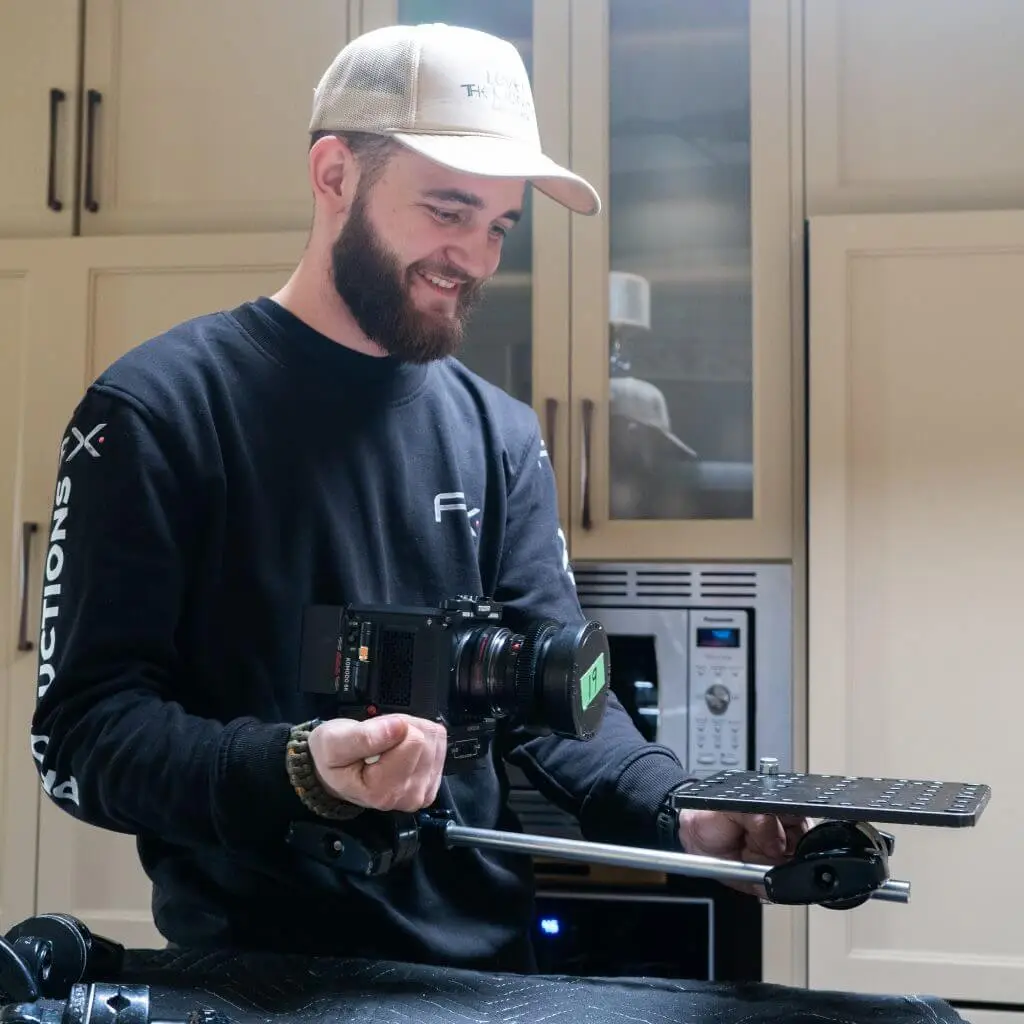 FX Productions Canada A man wearing a cap and dark sweatshirt holds a digital camera rig with a mounting plate in a kitchen setting, capturing behind-the-scenes moments for FX Productions Canada. Toronto Video Production Company