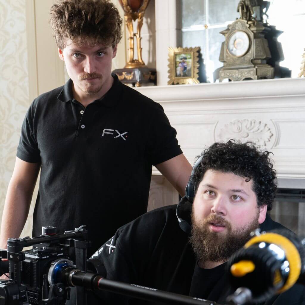 FX Productions Canada Two men working with camera equipment indoors for FX Productions Canada; one stands in a black polo shirt, the other sits with a headset and black shirt, in front of a fireplace with ornate decor. Toronto Video Production Company