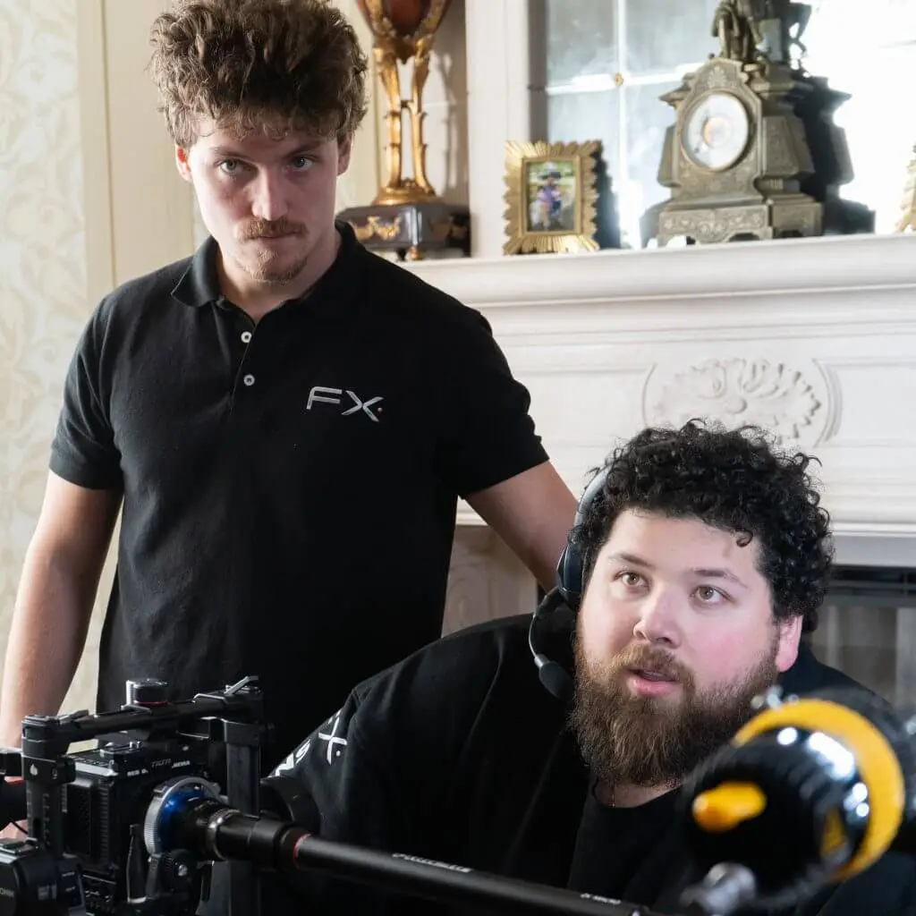FX Productions Canada Two men working with camera equipment indoors for FX Productions Canada; one stands in a black polo shirt, the other sits with a headset and black shirt, in front of a fireplace with ornate decor. Toronto Video Production Company