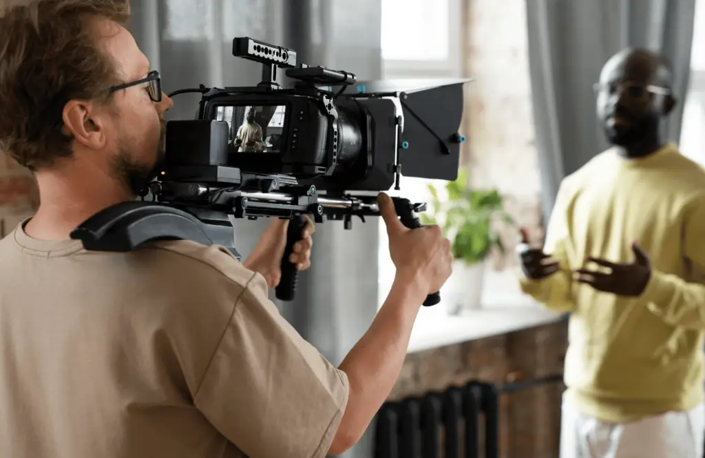 FX Productions Canada A person from FX Productions Canada operates a shoulder-mounted video camera, filming another person who is speaking and gesturing indoors near a window. Toronto Video Production Company