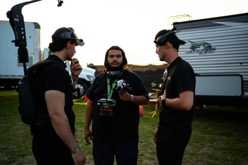 FX Productions Canada Three men in black attire stand outdoors—two with headsets, one holding a camera rig—conversing near a trailer and equipment, embodying the professionalism of a Toronto Video Production company. Toronto Video Production Company