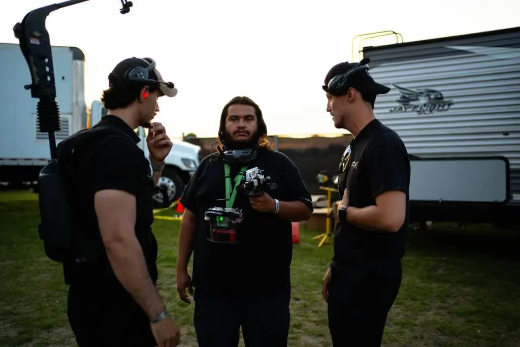 FX Productions Canada Three men in black attire stand outdoors—two with headsets, one holding a camera rig—conversing near a trailer and equipment, embodying the professionalism of a Toronto Video Production company. Toronto Video Production Company