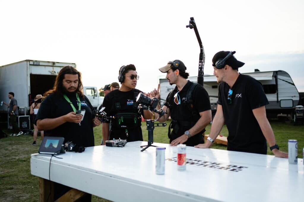 FX Productions Canada Four crew members in black shirts and headsets stand around a white table with film equipment, drinks, and gear—an energetic scene from an FX Productions Canada Toronto Video Production set, outdoors near trucks and a trailer. Toronto Video Production Company