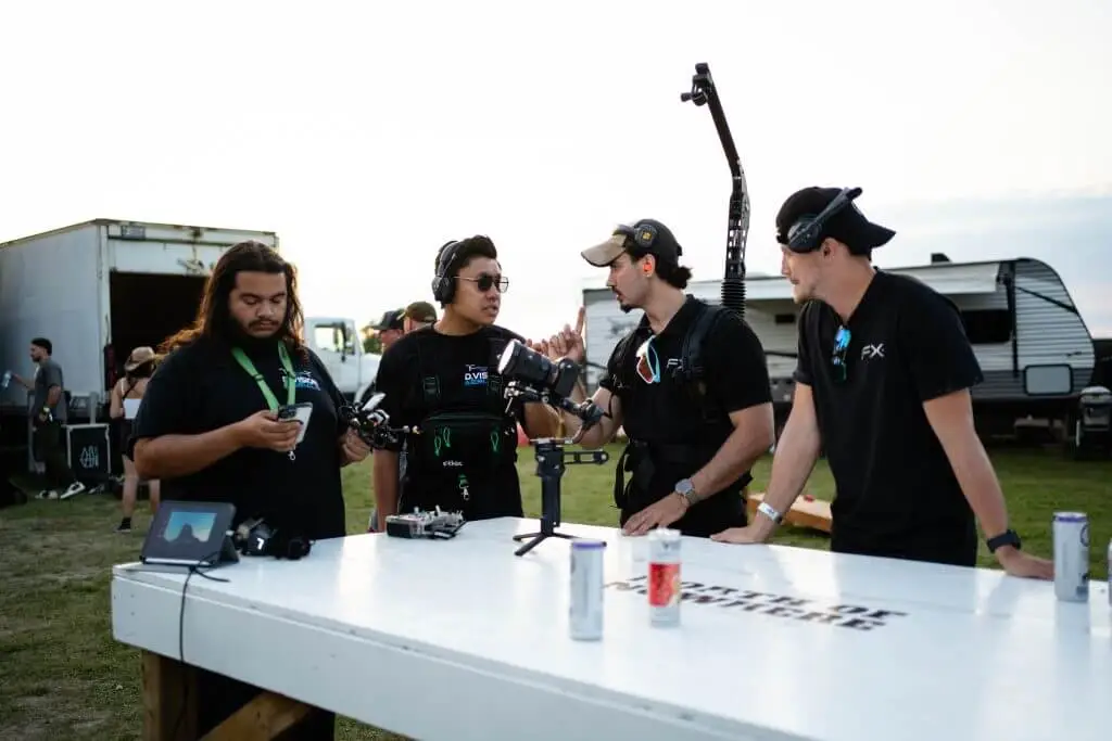 FX Productions Canada Four crew members in black shirts and headsets stand around a white table with film equipment, drinks, and gear—an energetic scene from an FX Productions Canada Toronto Video Production set, outdoors near trucks and a trailer. Toronto Video Production Company