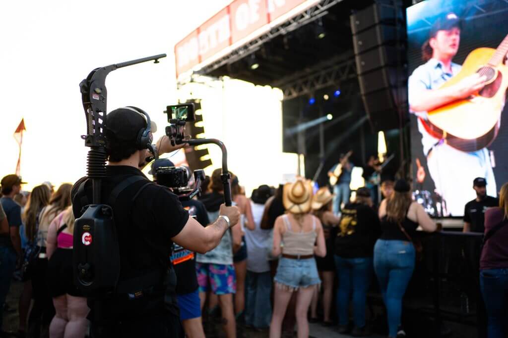 FX Productions Canada A cameraman from FX Productions Canada films a live outdoor concert as a crowd watches the stage, where a large screen displays a close-up of a musician playing guitar. Toronto Video Production Company