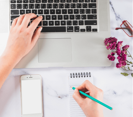 FX Productions Canada Person typing on a laptop and writing in a notepad, with a smartphone, dried flowers, and eyeglasses on a white desk—capturing the creative process at FX Productions Canada, a leading Toronto Video Production company. Toronto Video Production Company