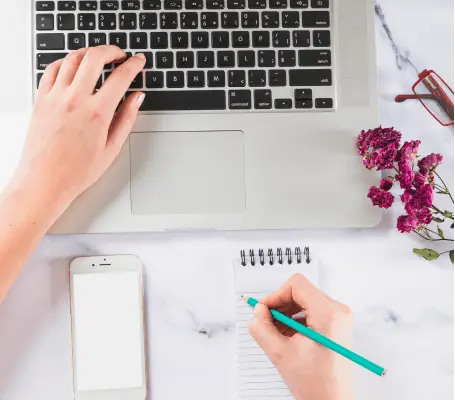 FX Productions Canada Person typing on a laptop and writing in a notepad, with a smartphone, dried flowers, and eyeglasses on a white desk—capturing the creative process at FX Productions Canada, a leading Toronto Video Production company. Toronto Video Production Company