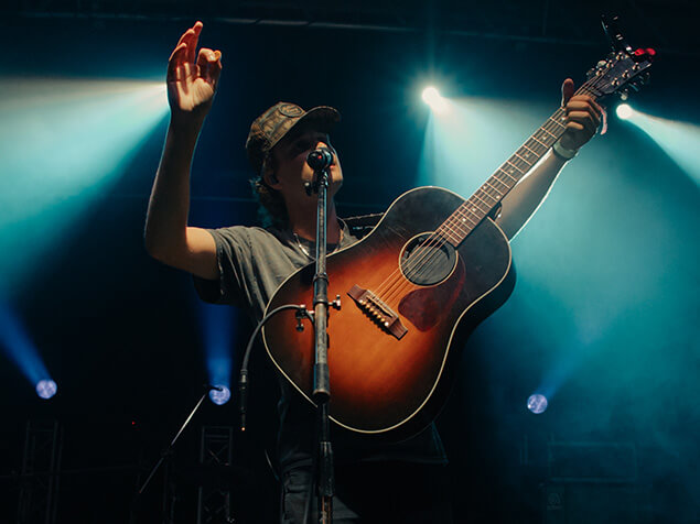 FX Productions Canada A person wearing a cap stands on stage with a guitar, one arm raised, singing or speaking into a microphone under stage lights—captured by Toronto Video Production professionals. Toronto Video Production Company