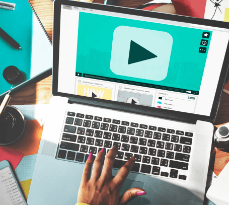 FX Productions Canada A person with pink nail polish uses a laptop displaying a large play button on a video player screen, surrounded by colorful office supplies—capturing the creative spirit of a Toronto Video Production company. Toronto Video Production Company