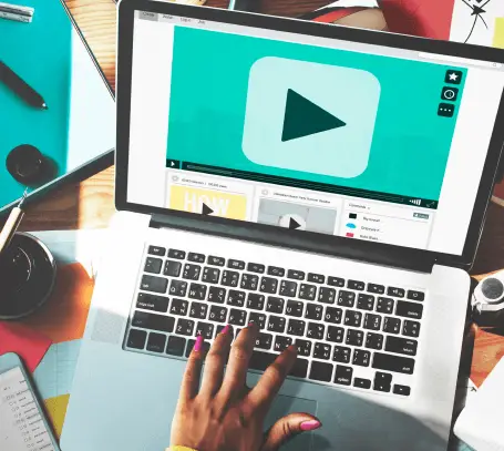 FX Productions Canada A person with pink nail polish uses a laptop displaying a large play button on a video player screen, surrounded by colorful office supplies—capturing the creative spirit of a Toronto Video Production company. Toronto Video Production Company
