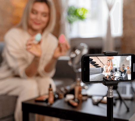 FX Productions Canada A woman sits on a couch holding two makeup sponges while recording a video with a smartphone on a tripod; makeup products are on the table in front of her, showcasing content created by Toronto Video Production experts. Toronto Video Production Company