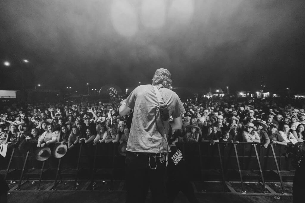 FX Productions Canada A musician stands onstage facing a large crowd at night, with stage lights above and the audience behind a barrier. Captured in black and white by FX Productions Canada, this image showcases expert Toronto Video Production. Toronto Video Production Company