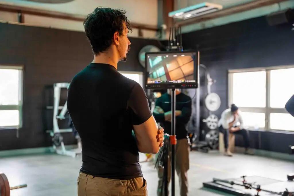 FX Productions Canada A man in a black shirt stands with arms crossed, looking at a monitor in a gym setting with equipment and large windows, capturing the behind-the-scenes vibe of a Toronto Video Production shoot. Toronto Video Production Company