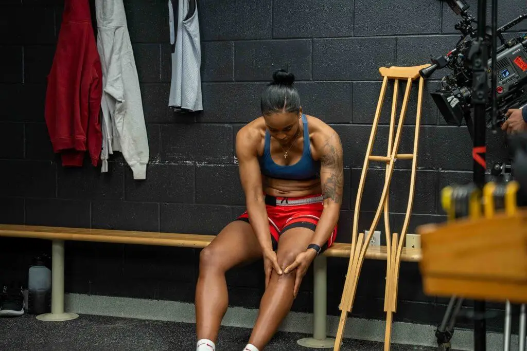 FX Productions Canada A woman in athletic wear sits on a bench in a locker room, touching her leg. Crutches and hanging clothes are nearby, with a Toronto Video Production crew filming her. Toronto Video Production Company