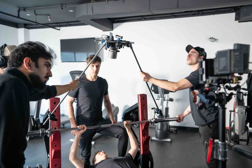 FX Productions Canada A film crew from FX Productions Canada records a person bench pressing weights in a gym, with one team member holding a boom microphone and another operating the camera. Toronto Video Production Company