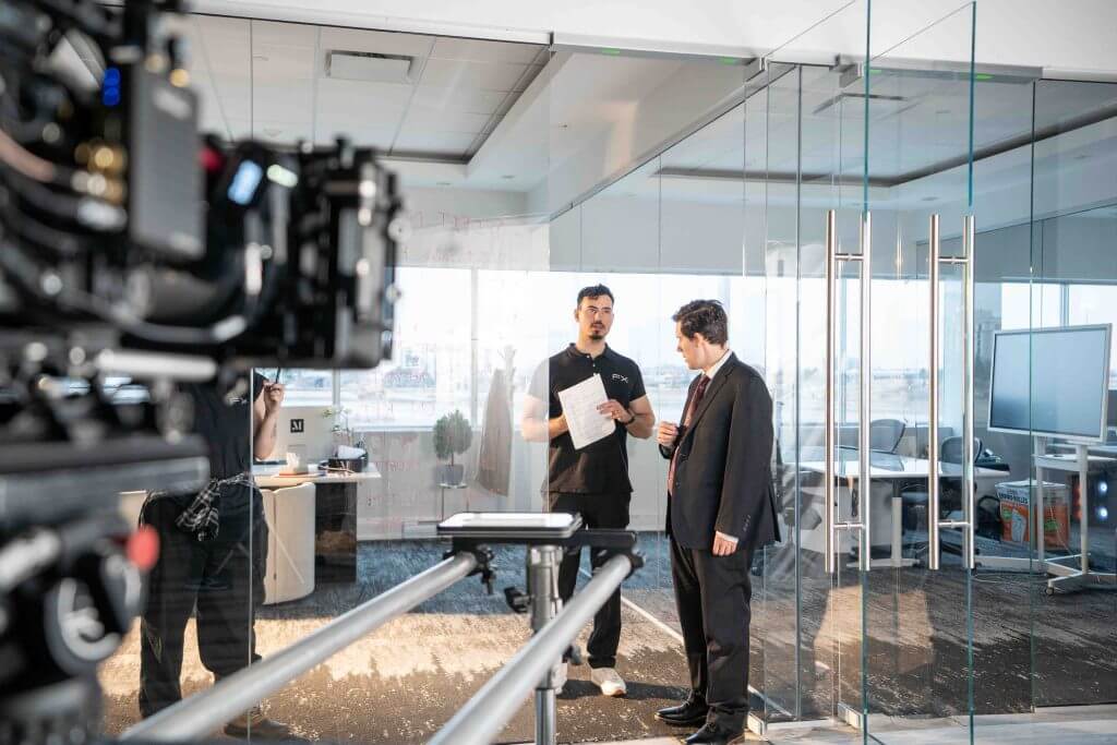 FX Productions Canada Two men stand talking in a modern glass-walled office while a camera on a track films them; one man holds papers, and crew members from FX Productions Canada are visible in the background. Toronto Video Production Company