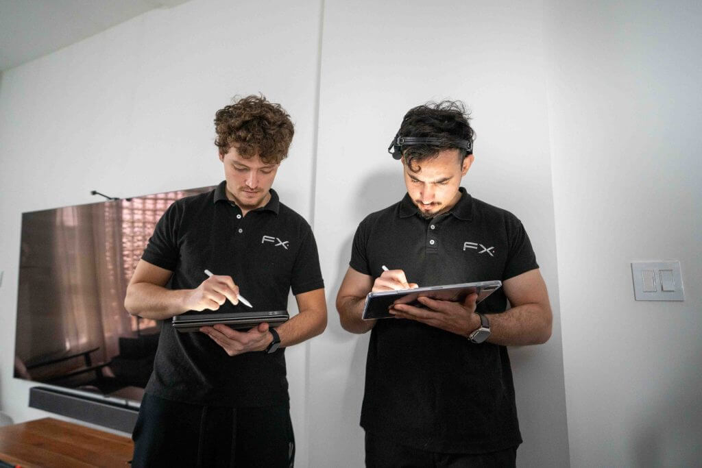 FX Productions Canada Two men in matching black FX Productions Canada polo shirts use styluses to write on tablets while standing indoors next to a wall and a flat-screen TV. Toronto Video Production Company