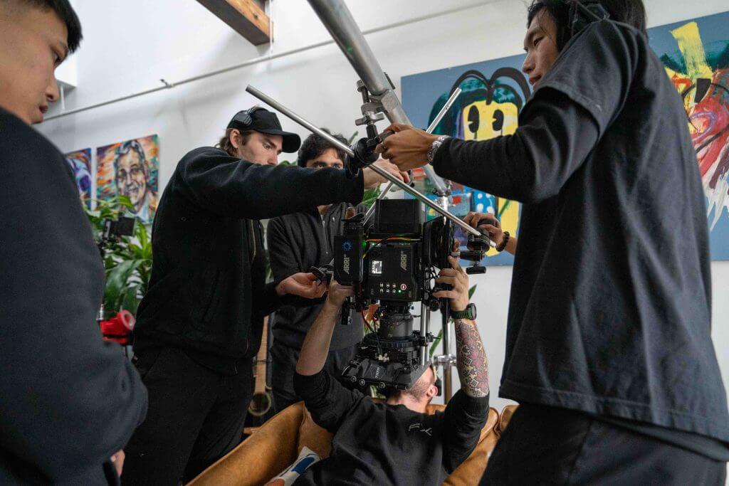 FX Productions Canada A film crew from a Toronto Video Production company adjusts camera equipment on set indoors, with one member lying down holding the camera and others assisting. Paintings can be seen in the background. Toronto Video Production Company