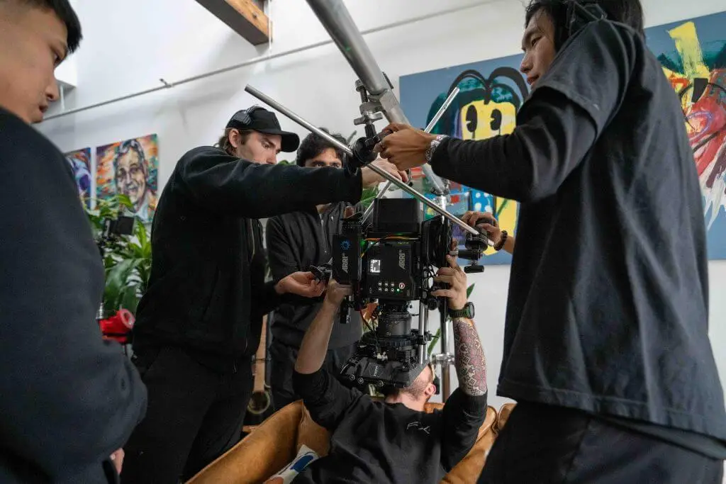 FX Productions Canada A film crew from a Toronto Video Production company adjusts camera equipment on set indoors, with one member lying down holding the camera and others assisting. Paintings can be seen in the background. Toronto Video Production Company
