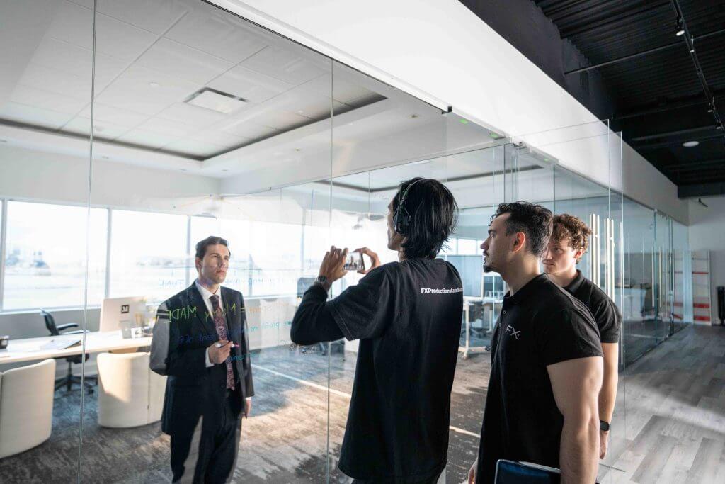 FX Productions Canada A man in a suit stands in a modern office while three casually dressed men face him, one holding a phone as if recording—a scene that captures the energy of a Toronto Video Production team at work. Toronto Video Production Company
