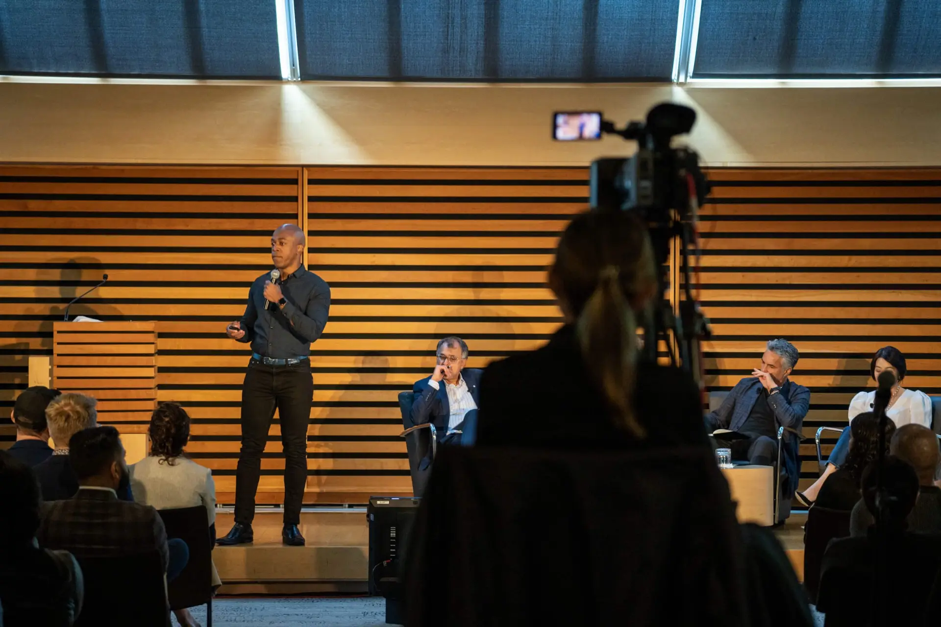 FX Productions Canada A speaker presents to an audience in a modern conference room, while a camera from FX Productions Canada records the event and panelists sit on stage in the background. Toronto Video Production Company