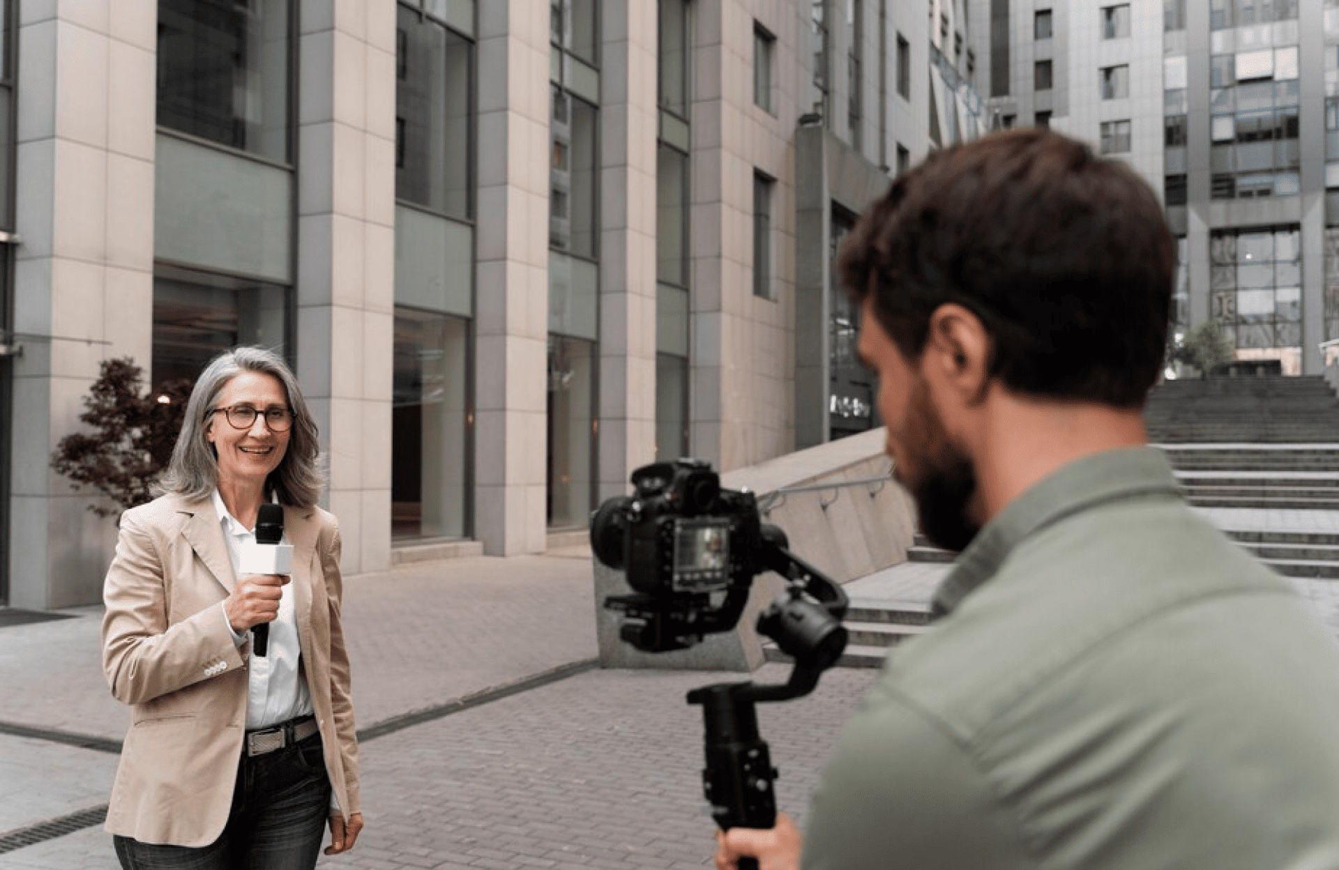 FX Productions Canada A woman holding a microphone stands outside in front of tall buildings while a man from Toronto Production Company films her with a camera on a stabilizer. Toronto Video Production Company