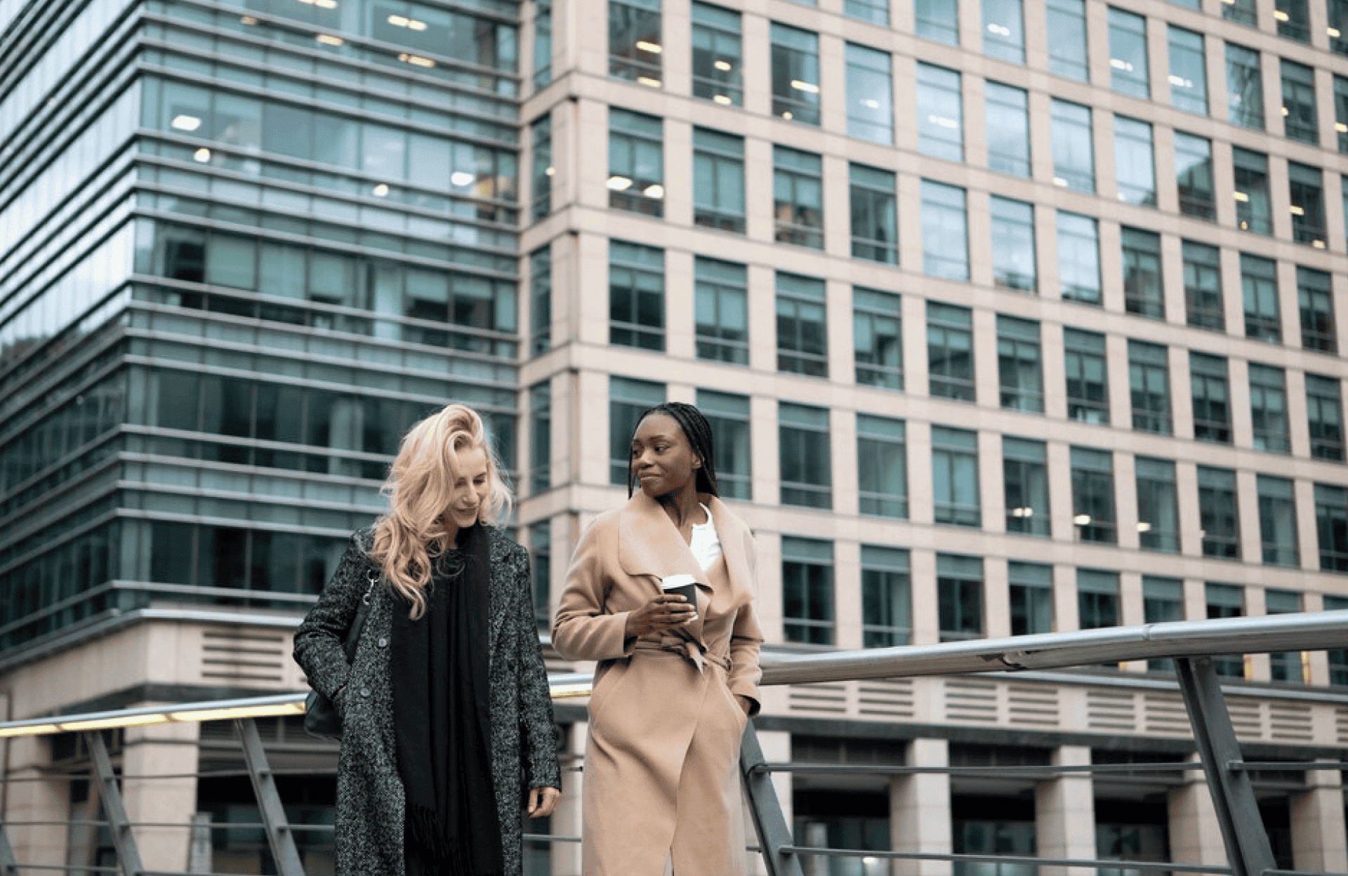 FX Productions Canada Two women in coats walk and talk on a modern pedestrian bridge, with a large glass office building in the background. One holds a coffee cup, reflecting the dynamic urban energy of FX Productions Canada, a top Toronto production company. Toronto Video Production Company