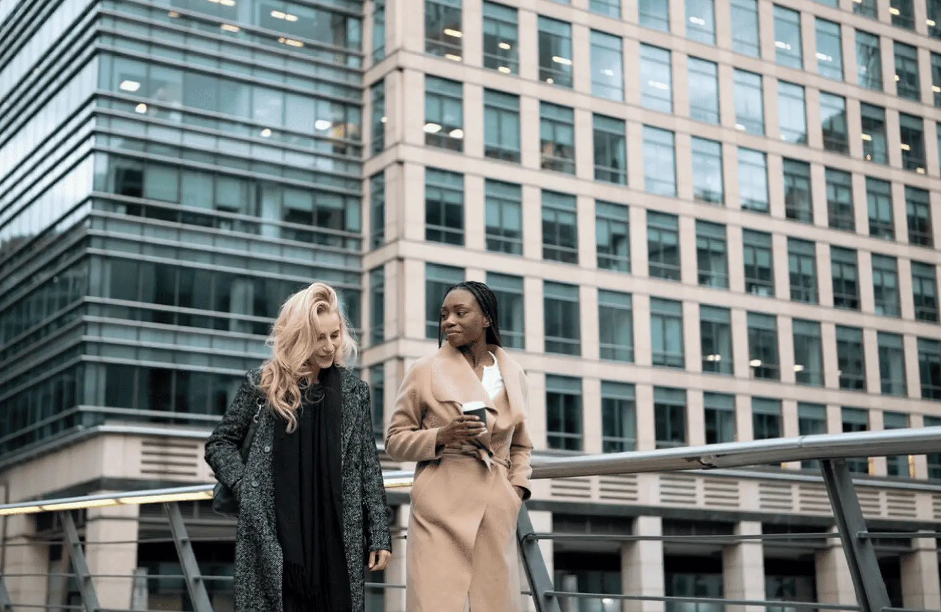 FX Productions Canada Two women in coats walk and talk on a modern pedestrian bridge, with a large glass office building in the background. One holds a coffee cup, reflecting the dynamic urban energy of FX Productions Canada, a top Toronto production company. Toronto Video Production Company