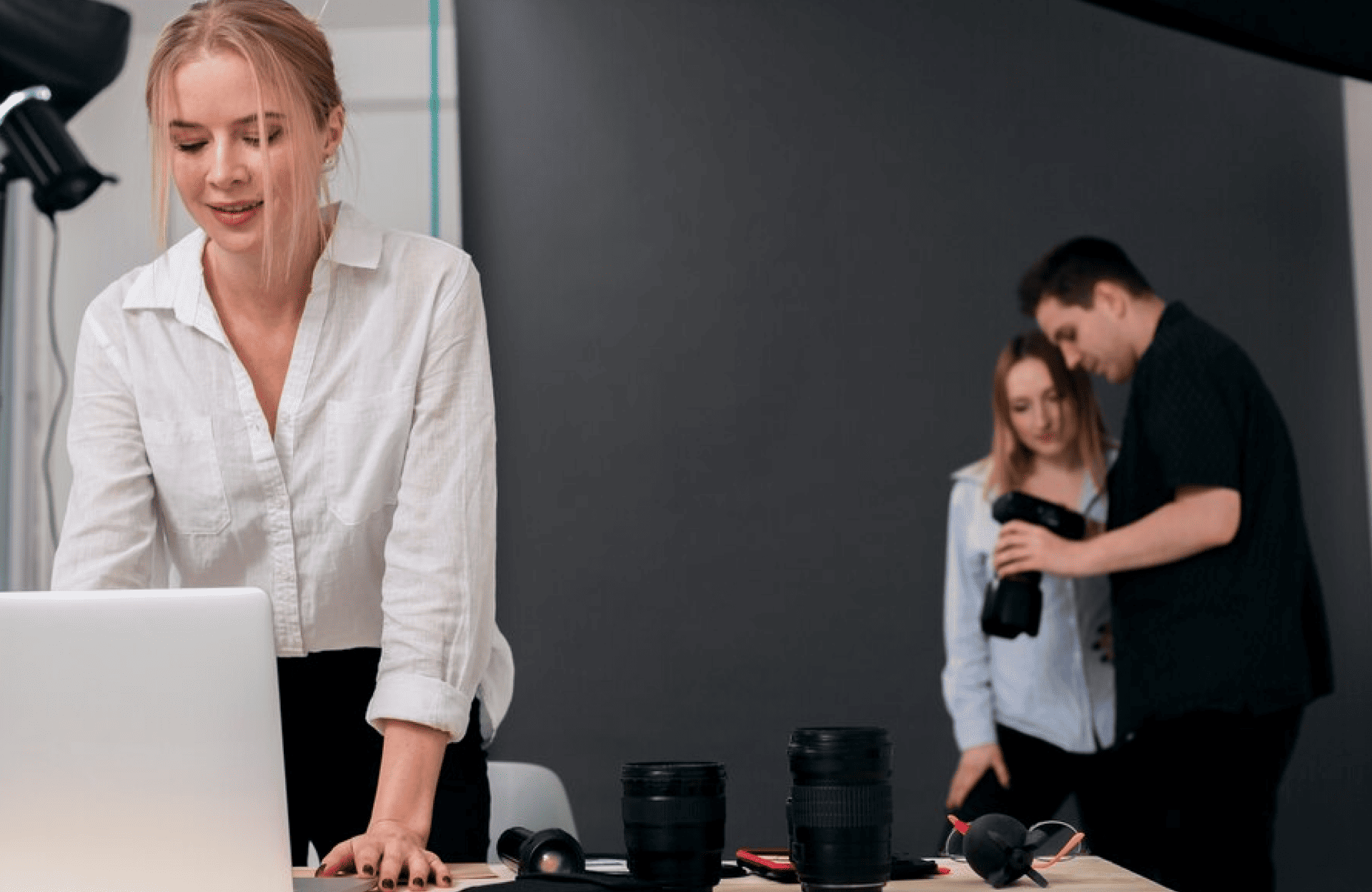 FX Productions Canada A woman works at a laptop on a desk with camera lenses, while two people in the background from FX Productions Canada examine a camera against a dark backdrop. Toronto Video Production Company