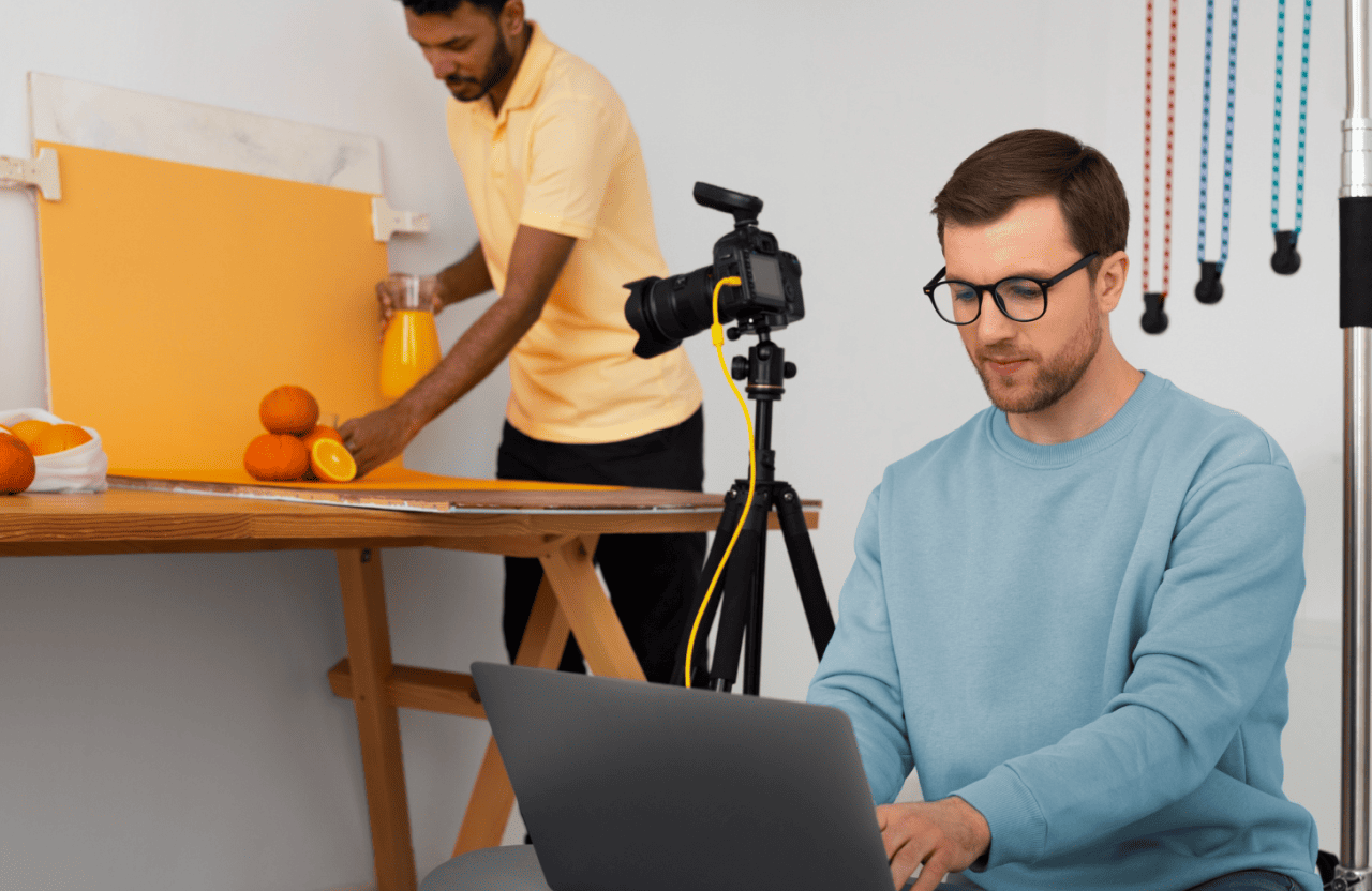 FX Productions Canada Two men work in a studio; one arranges oranges and juice by a camera on a tripod, while the other types on a laptop—capturing the creative process at FX Productions Canada. Toronto Video Production Company