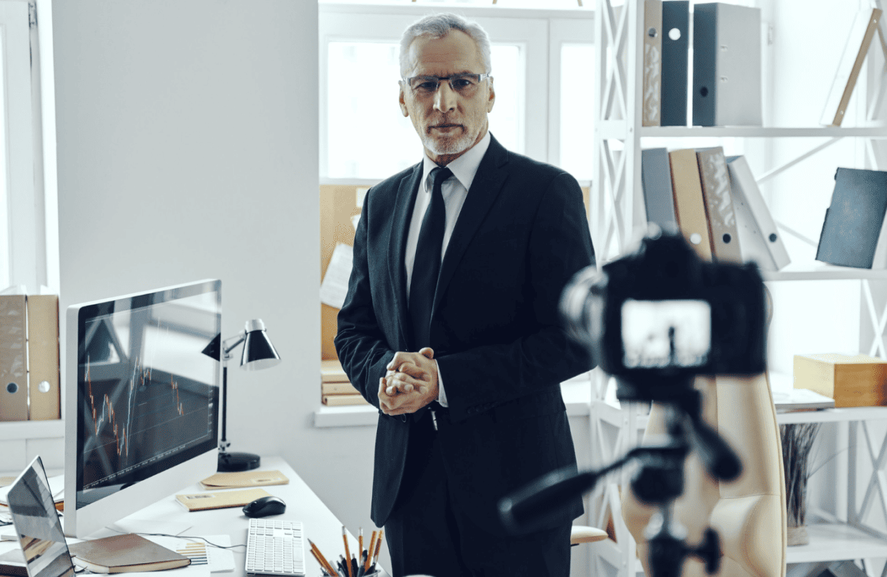 FX Productions Canada A man in a suit stands in an office with shelves and a computer, facing a camera on a tripod as if recording or live streaming for FX Productions Canada, a Toronto Production Company. Toronto Video Production Company