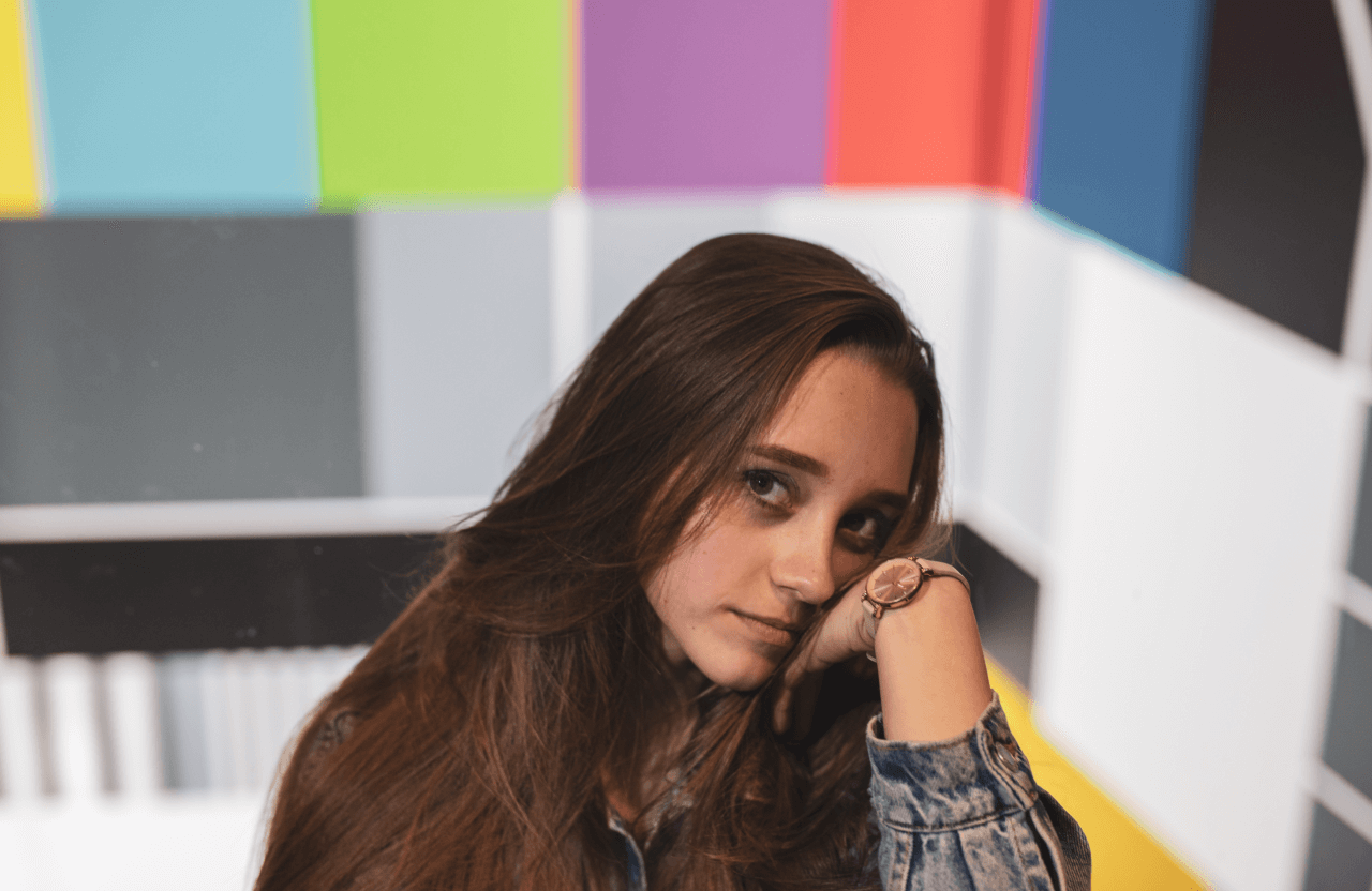 FX Productions Canada A young woman with long brown hair rests her head on her hand, sitting in front of a colorful, geometric background designed by FX Productions Canada. Toronto Video Production Company