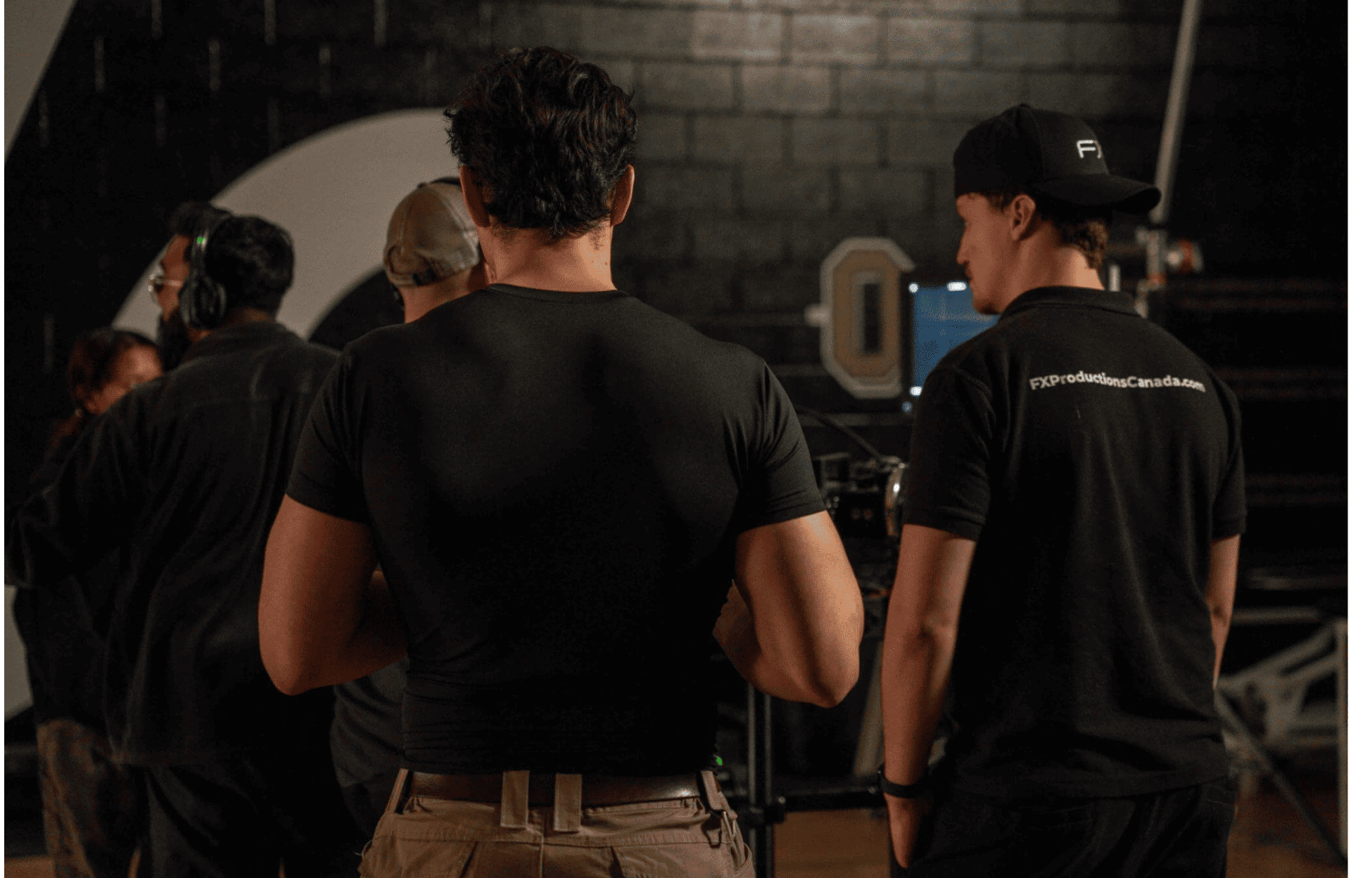 FX Productions Canada A group of people stand indoors, facing away from the camera, with two men in the foreground wearing black shirts and one holding a cap—capturing a behind-the-scenes moment at an FX Productions Canada event. Toronto Video Production Company