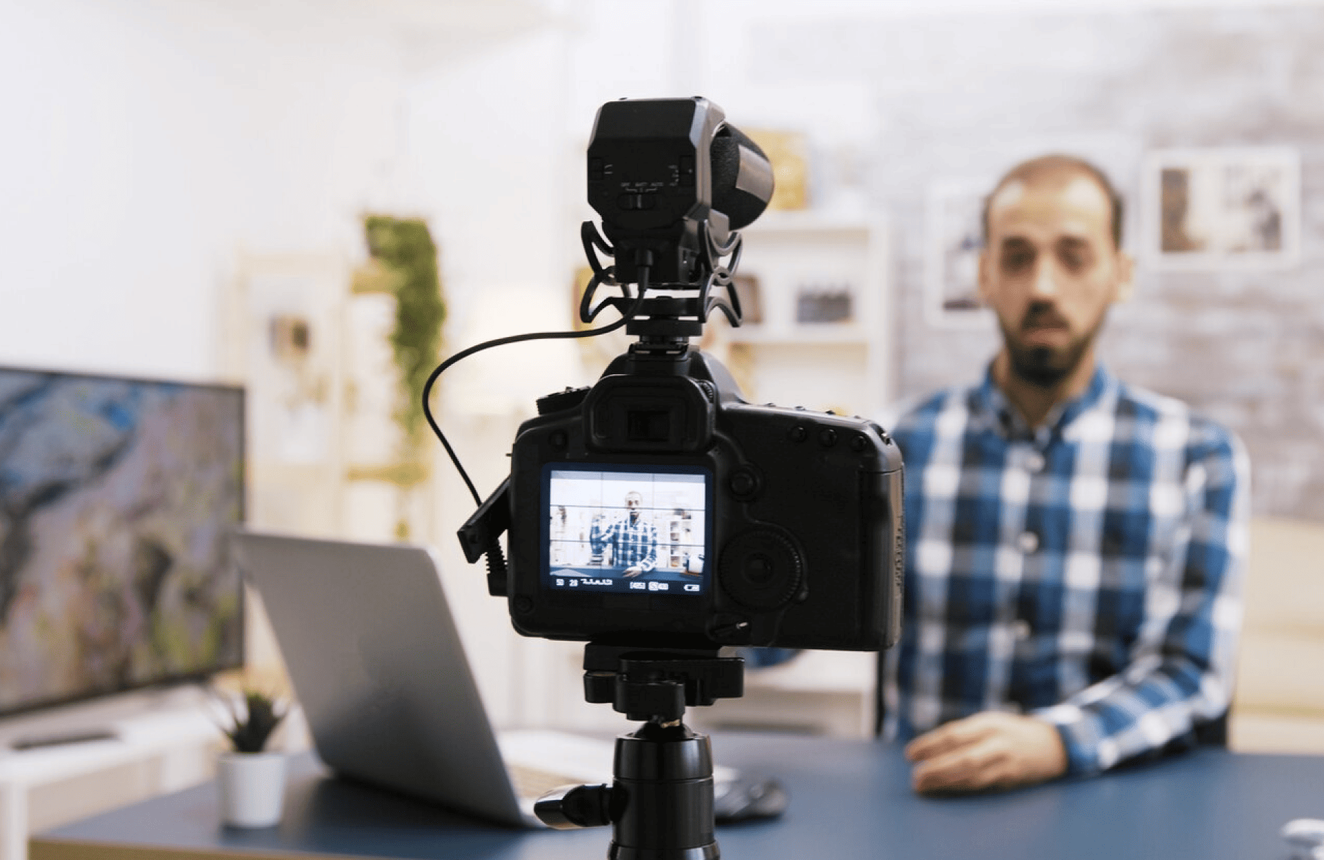 FX Productions Canada A camera on a tripod records a man in a blue plaid shirt sitting at a desk with a laptop and plant in the background. Toronto Video Production Company