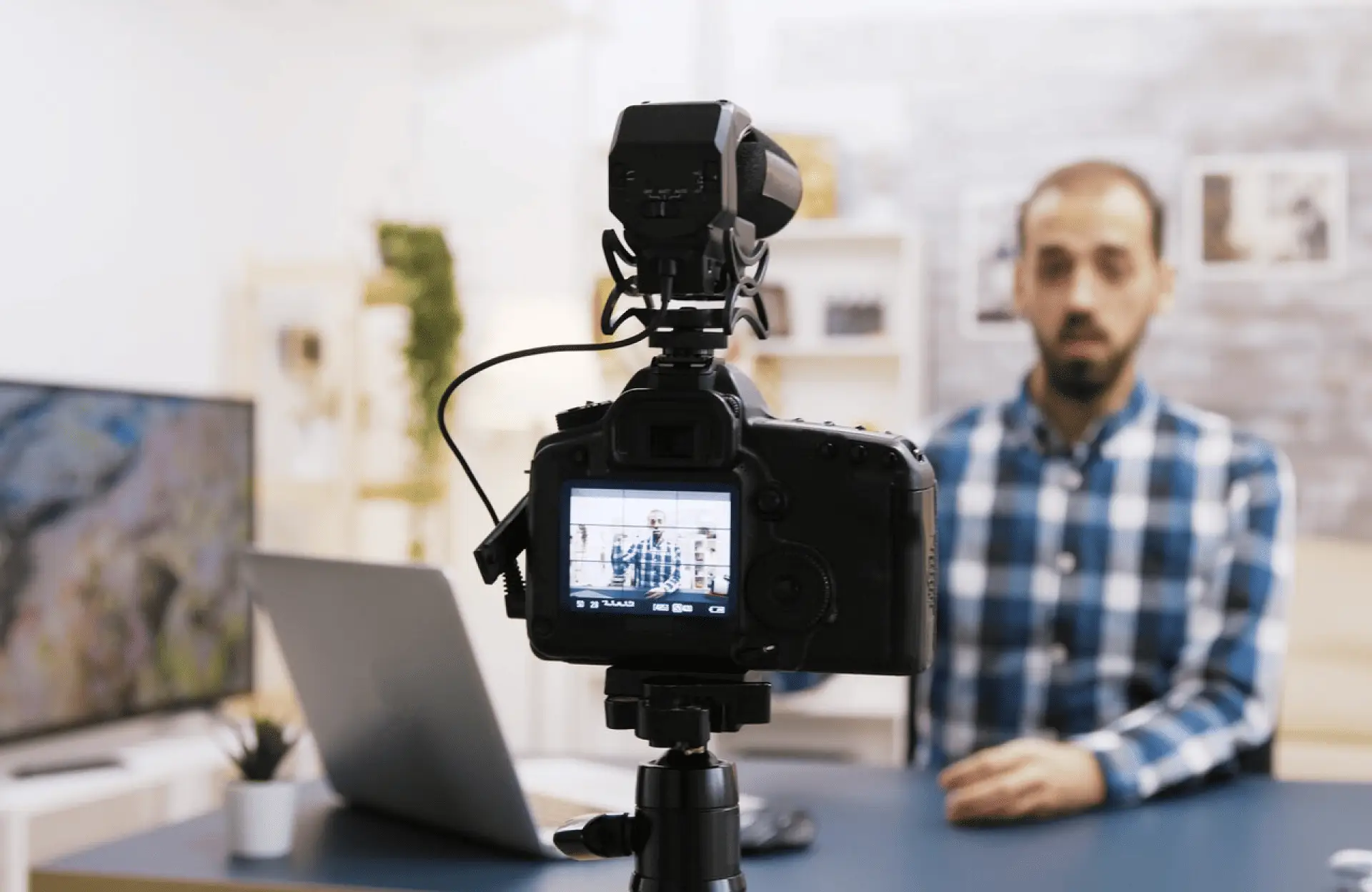 FX Productions Canada A camera on a tripod records a man in a blue plaid shirt sitting at a desk with a laptop and plant in the background. Toronto Video Production Company