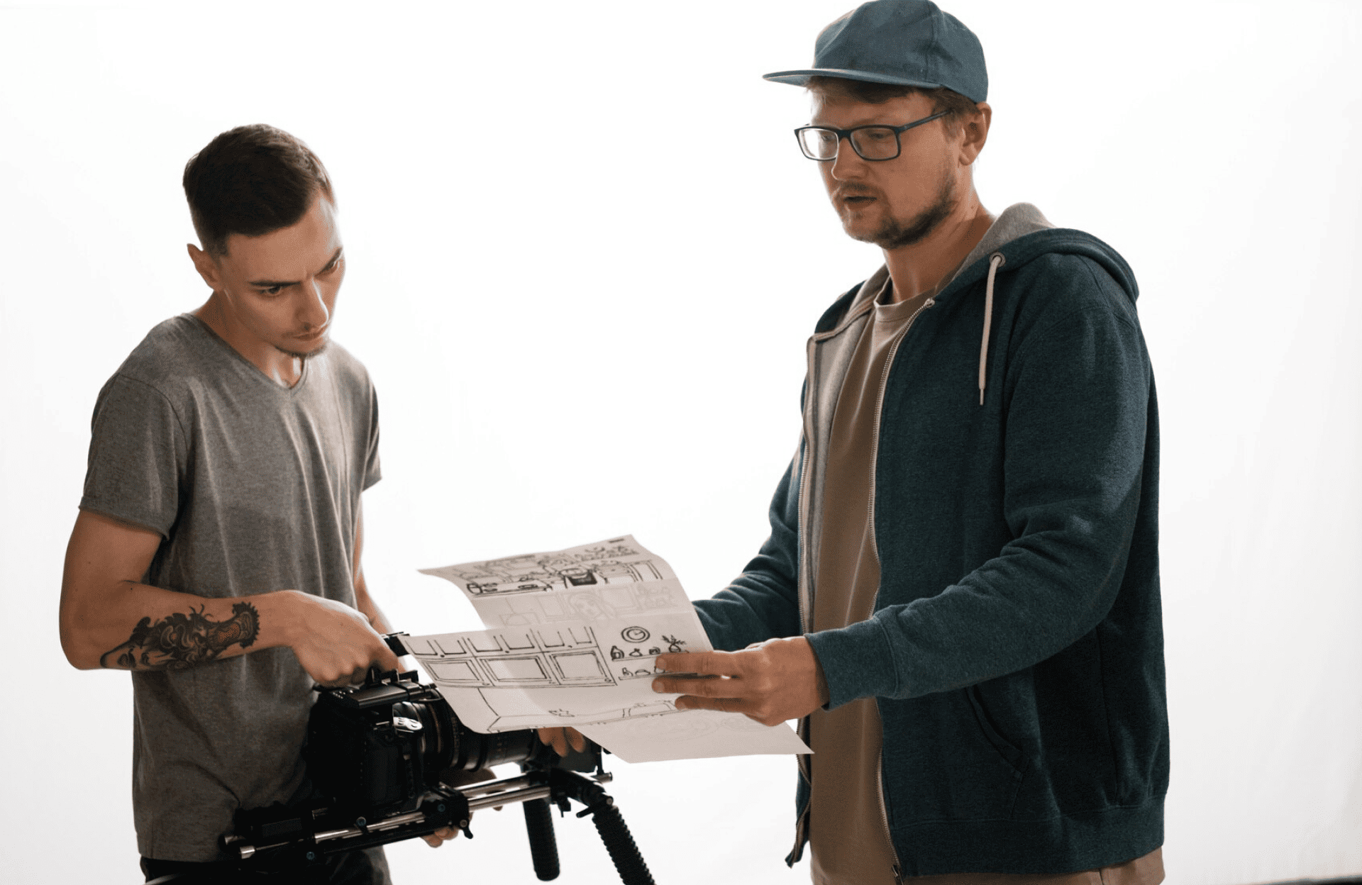 FX Productions Canada Two men stand together; one adjusts a camera on a tripod while the other holds and examines a storyboard against a plain white background. Toronto Video Production Company