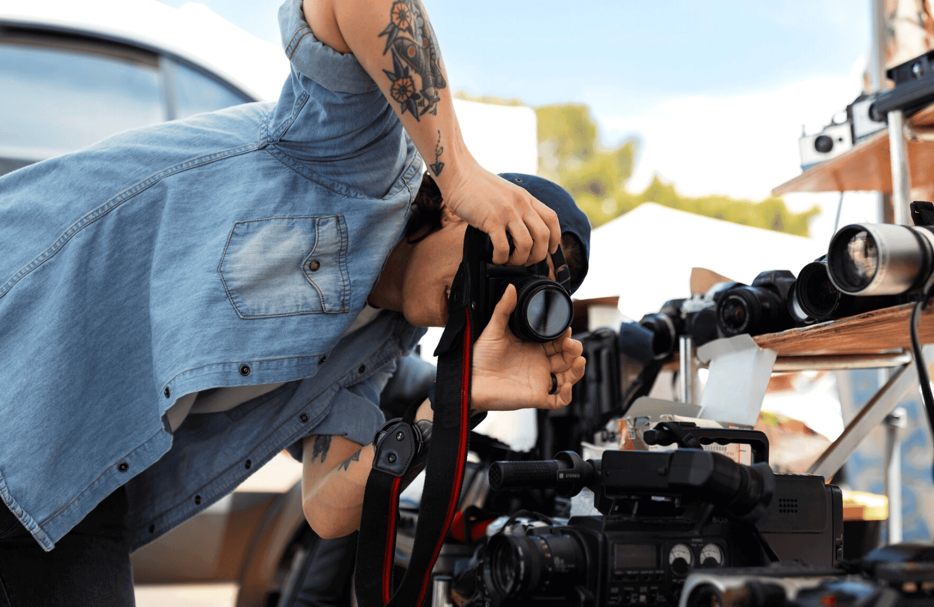FX Productions Canada A person in a denim shirt and tattoos takes a photo with a camera, surrounded by various cameras and lenses on a table at an outdoor market. Toronto Video Production Company