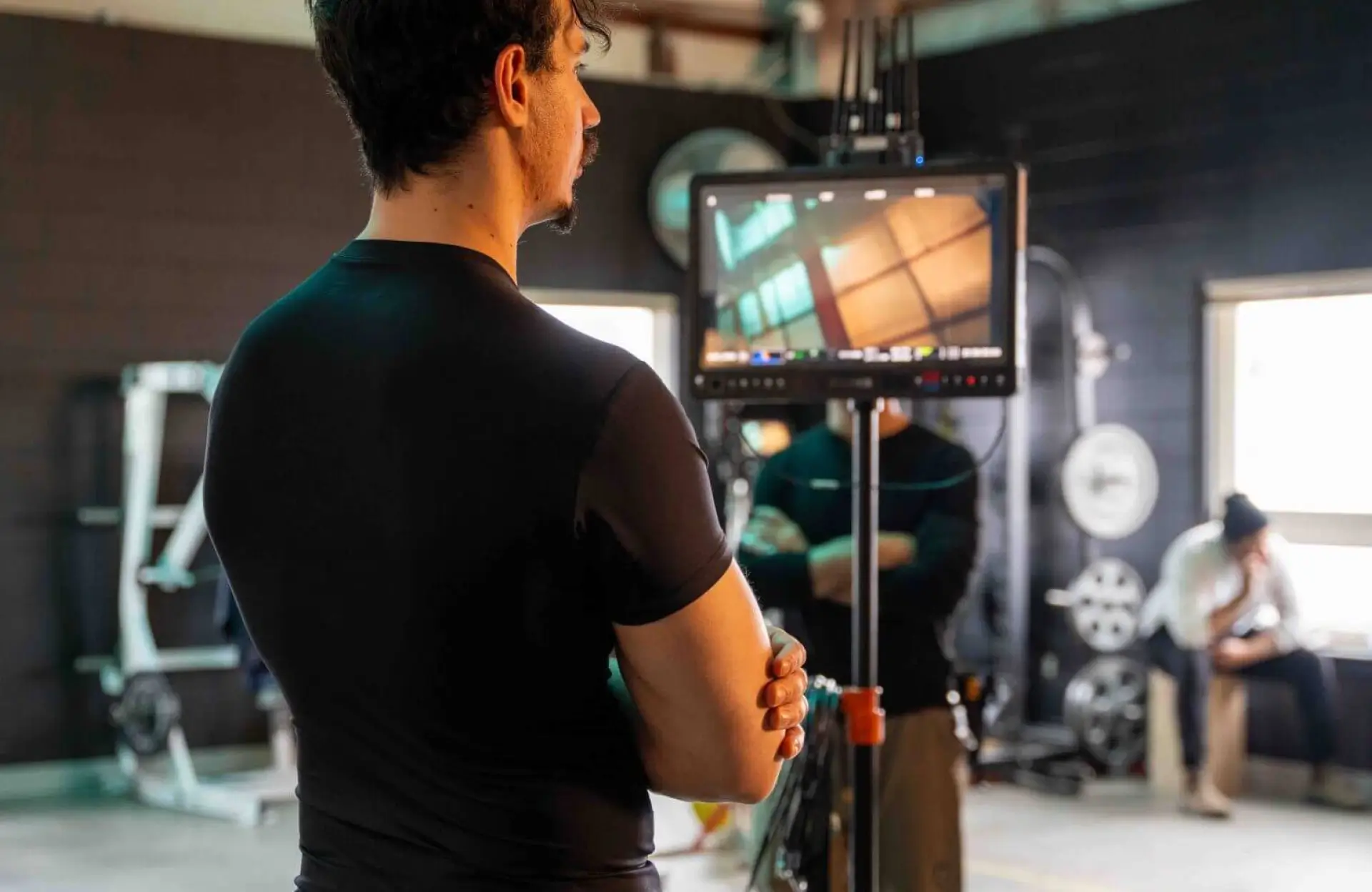 FX Productions Canada A person in a black shirt stands with arms crossed in front of a monitor displaying a building interior, inside a gym with weights and fitness equipment. Toronto Video Production Company