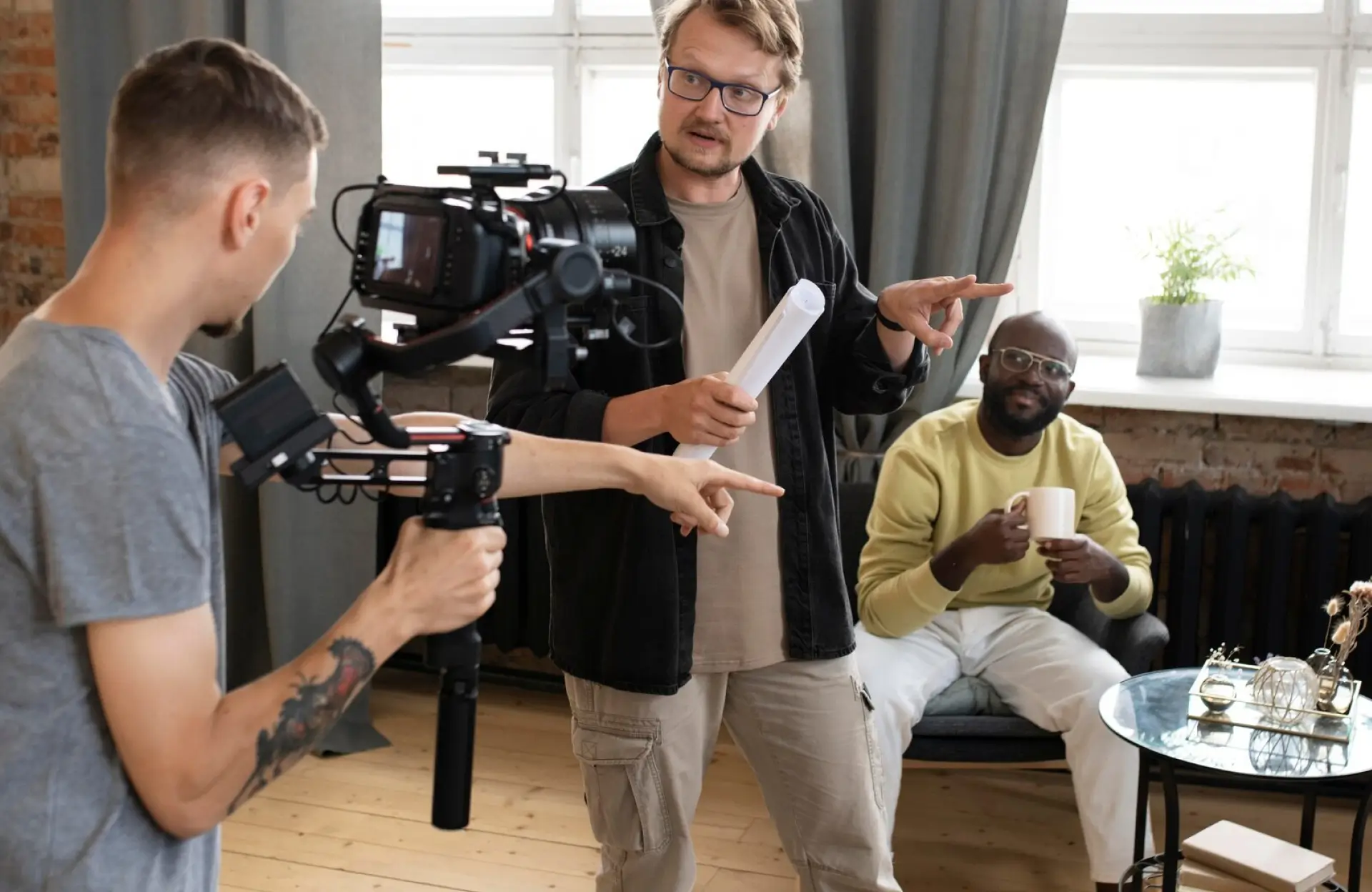 FX Productions Canada Three men are filming indoors; one holds a camera, another gestures with papers, and the third sits on a sofa holding a cup. Toronto Video Production Company