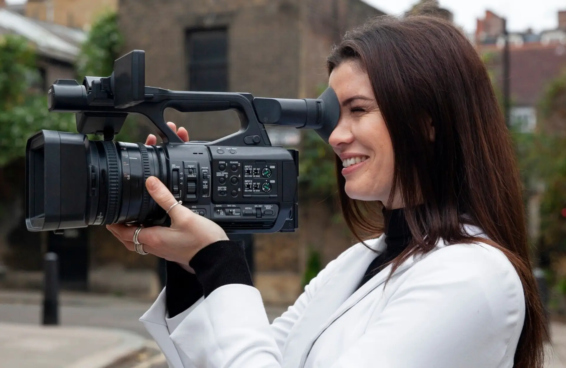 FX Productions Canada A woman wearing a white jacket holds a large video camera and looks through the viewfinder while standing outdoors. Toronto Video Production Company