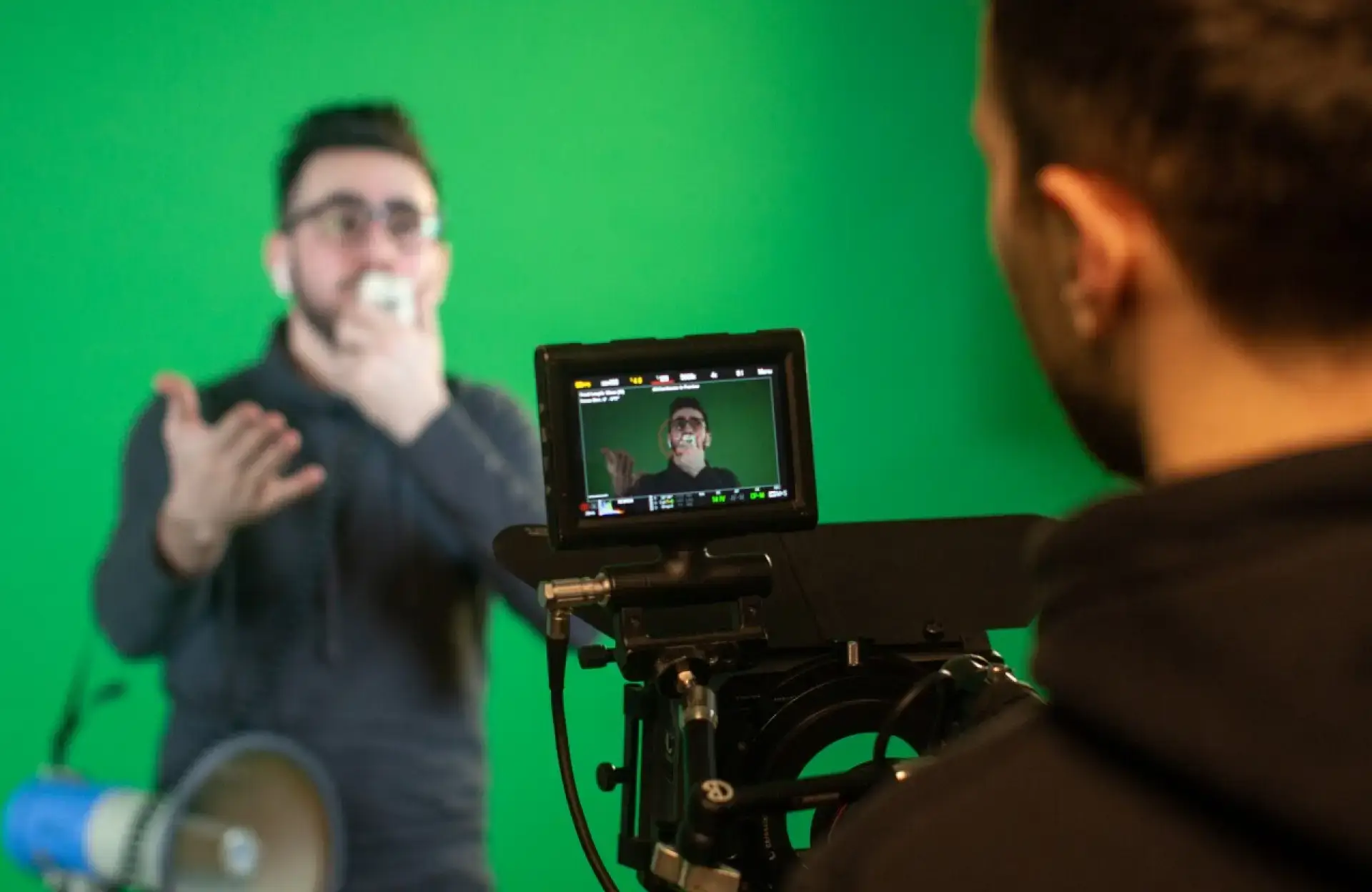 FX Productions Canada A person records another individual in front of a green screen. The camera monitor displays the subject, who is gesturing with one hand and holding an object near his mouth. Toronto Video Production Company