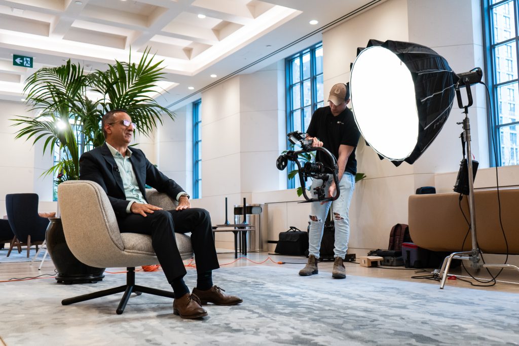 FX Productions Canada A man in a suit sits in an armchair for an interview while a cameraman films him with professional lighting in a modern office space. Toronto Video Production Company