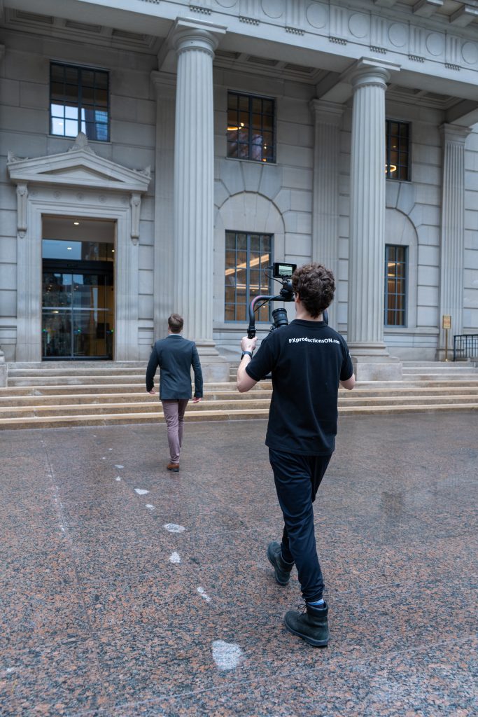 FX Productions Canada A person with a camera follows another person walking up the steps toward a large building with tall columns. Toronto Video Production Company