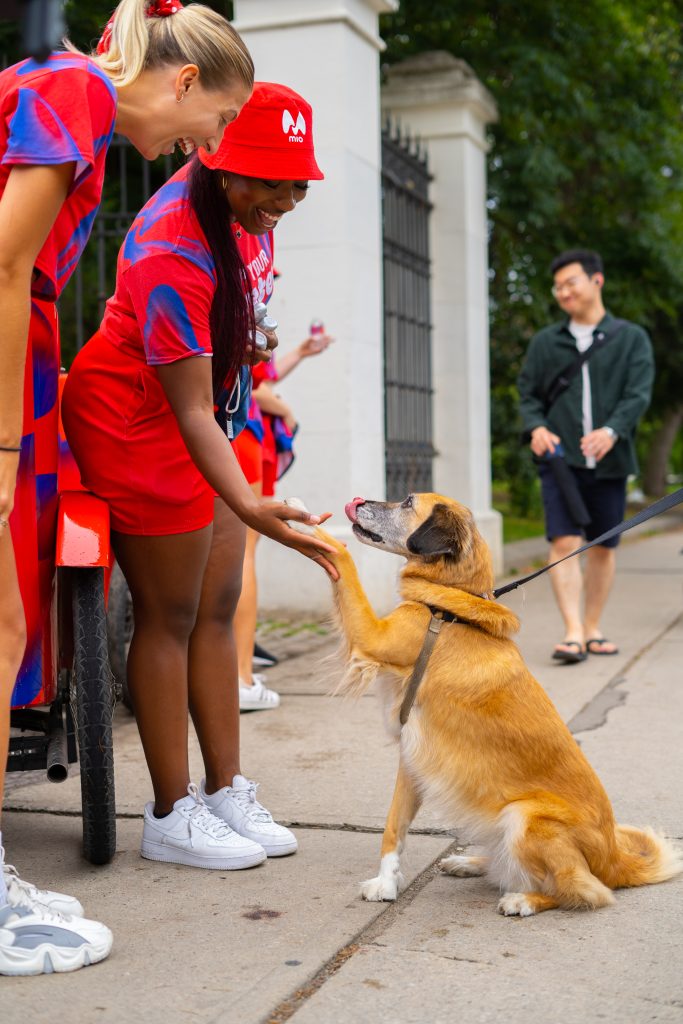 FX Productions Canada Two women in matching red and blue outfits kneel to shake hands with a brown and white dog, while a man walks by in the background. Toronto Video Production Company