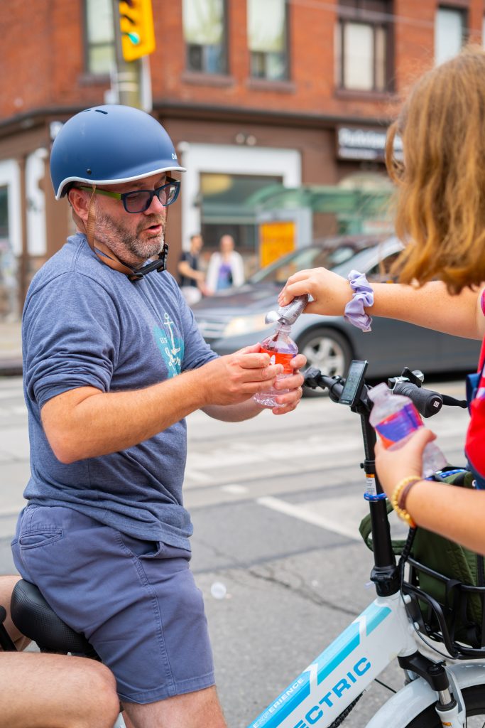 FX Productions Canada A man wearing a helmet sits on an electric bike while a girl hands him a bottle of water on a city street. Toronto Video Production Company