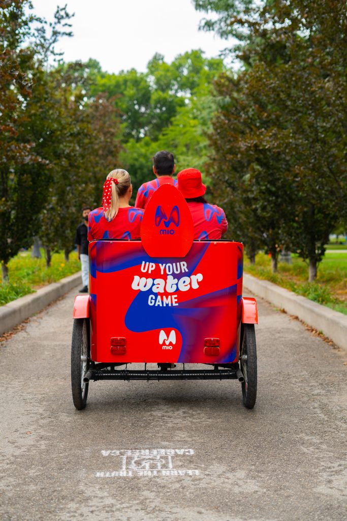 FX Productions Canada Three people in matching red and blue outfits ride a branded pedicab with "UP YOUR WATER GAME" text, on a paved path lined with trees. Toronto Video Production Company
