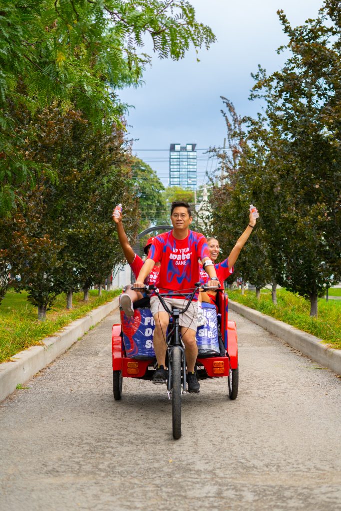 FX Productions Canada A man pedals a red tricycle with two passengers on a tree-lined path; all three wear matching red jerseys and hold up drinks. Toronto Video Production Company