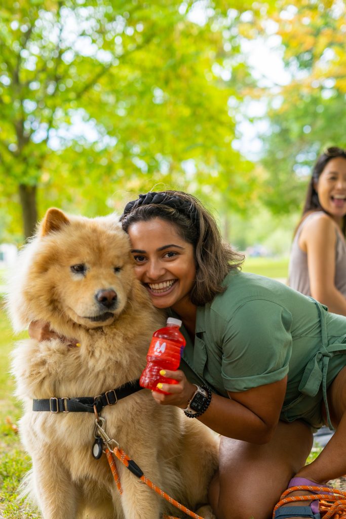 FX Productions Canada A woman crouches next to a fluffy dog, smiling and holding a red toy, while another person sits in the background in a park setting. Toronto Video Production Company